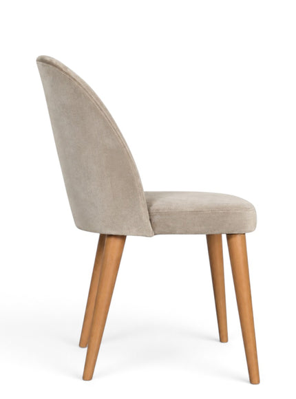 A modern dining chair with a curved back upholstered in light beige velvet, supported by four tapered natural wood legs, shown from a three-quarter angle on a white background.