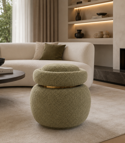 A round, low stool with a textured, bouclé-like fabric in a muted sage green, featuring a gold metallic band around its upper tier. The stool is placed in a modern living room setting on a beige rug in front of a cream-colored sofa.
