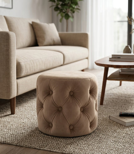 A round tufted ottoman stool in a warm taupe-beige velvet fabric, styled in a modern living room setting next to a beige sofa and a wooden coffee table.