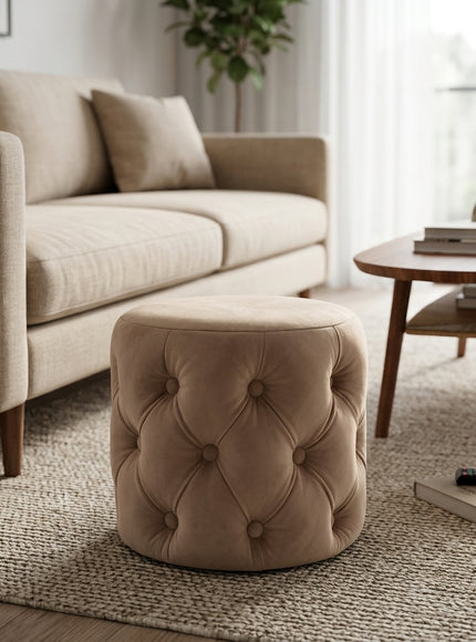 A round tufted ottoman stool in a warm taupe-beige velvet fabric, styled in a modern living room setting next to a beige sofa and a wooden coffee table.