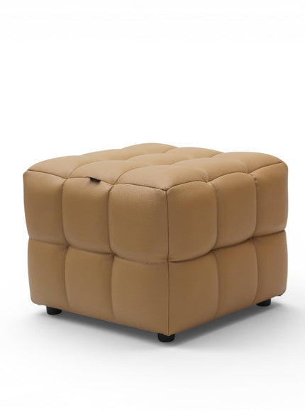 A square tufted ottoman in tan leather with a small top lift handle, standing on small black feet against a white background.