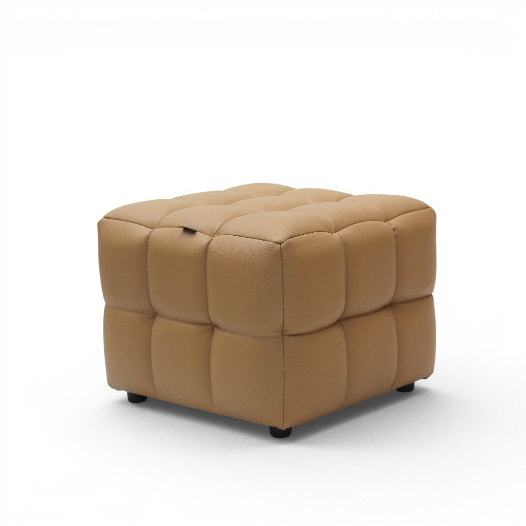 A square tufted ottoman in tan leather with a small top lift handle, standing on small black feet against a white background.