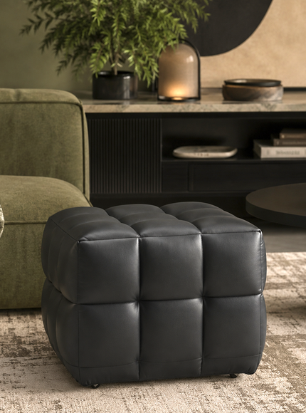 A black tufted cube ottoman with a padded leather-like finish, styled in a modern living room setting next to an olive green sofa and a round black coffee table.