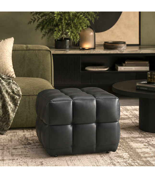A black tufted cube ottoman with a padded leather-like finish, styled in a modern living room setting next to an olive green sofa and a round black coffee table.