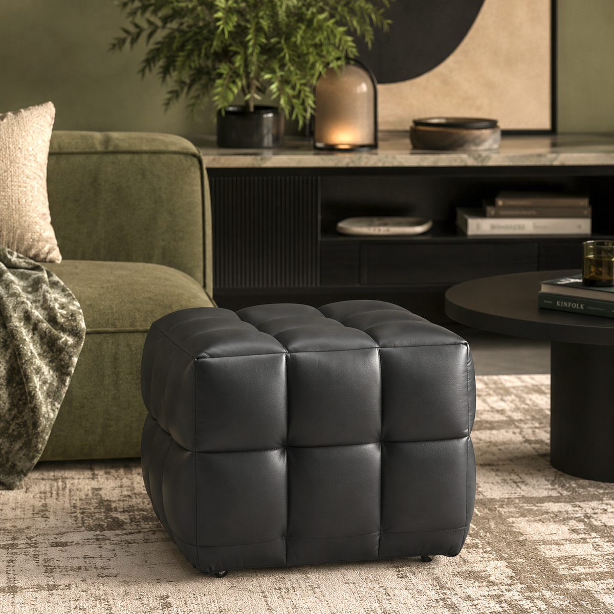 A black tufted cube ottoman with a padded leather-like finish, styled in a modern living room setting next to an olive green sofa and a round black coffee table.