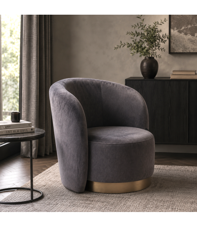 A modern barrel-style accent chair upholstered in dark gray velvet with a brushed gold metal base, set in a styled living room interior.