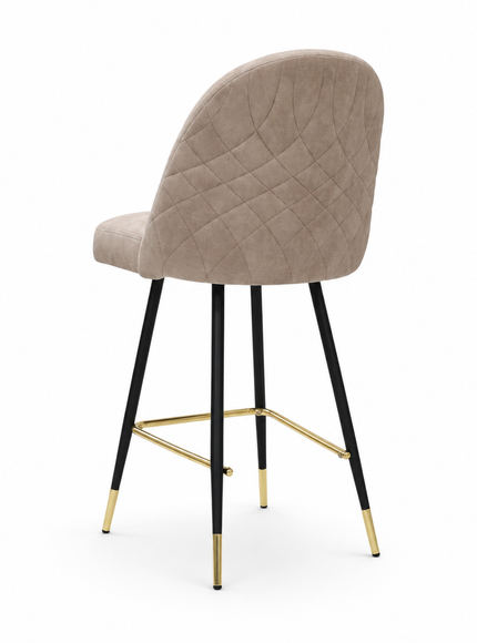 A modern bar stool with a rounded, upholstered seat and backrest in a taupe-beige fabric, supported by slim black metal legs accented with gold hardware and gold-colored foot caps, set against a plain white background.