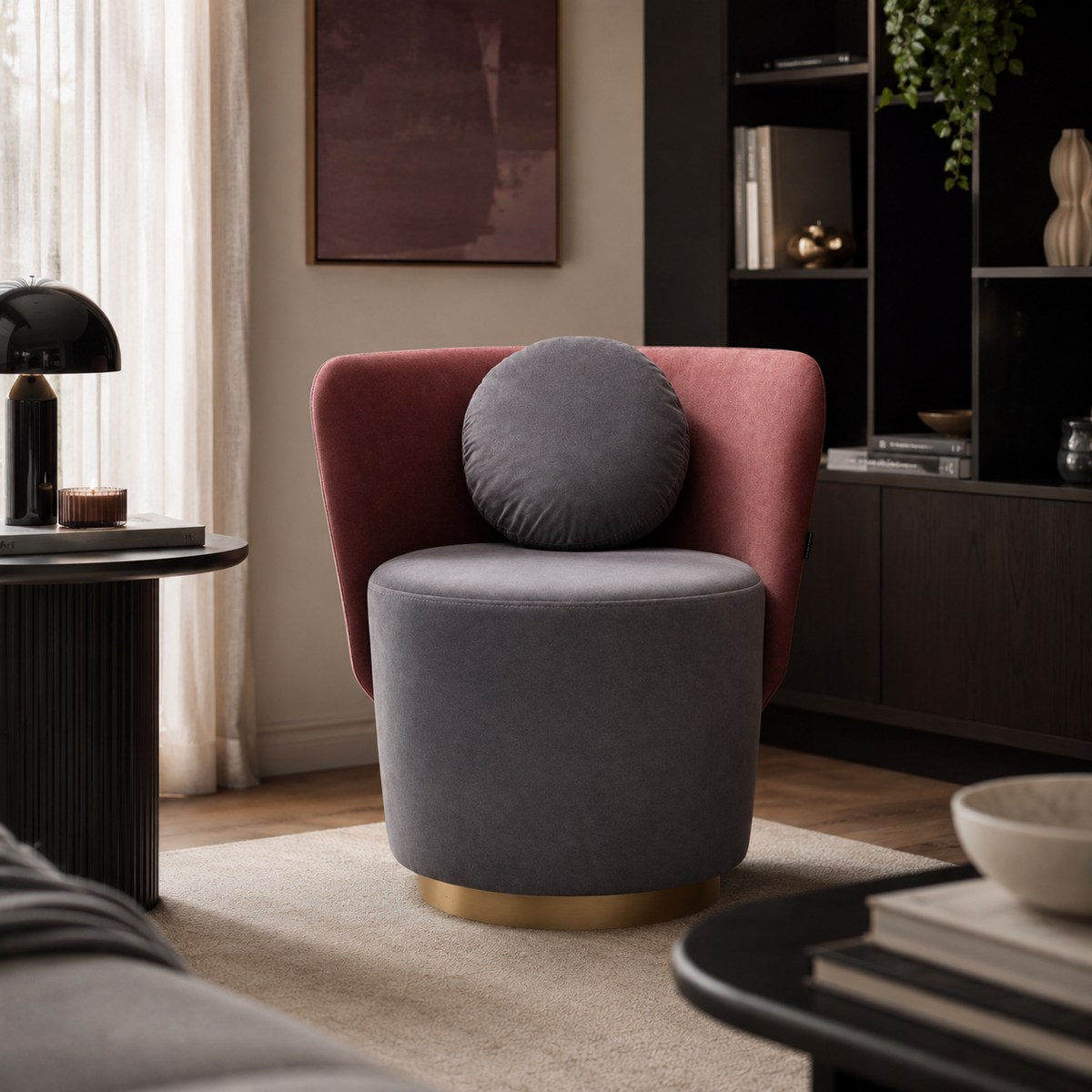 A modern accent chair in a styled living room, featuring a dusty rose velvet armchair with a round gray lumbar pillow and a cylindrical gray velvet seat on a gold-finished base.