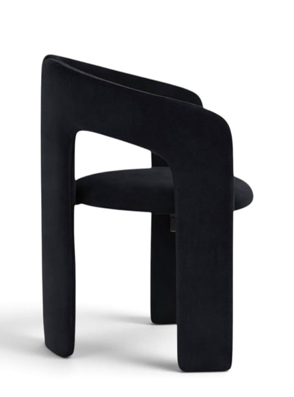 A modern black velvet dining chair with a unique split backrest and wide block legs, shown from the side on a white background.