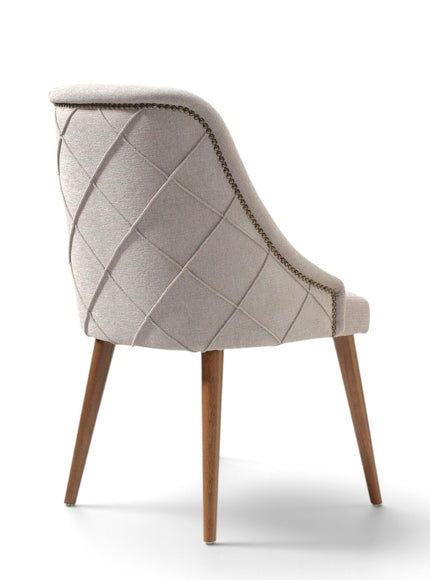 A light beige upholstered dining chair with a diamond-quilted pattern on the backrest, wooden legs, and brass nailhead trim, shown from a three-quarters angle on a white background.