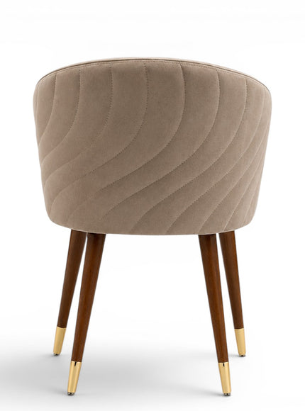 A modern barrel-style dining chair with a rounded, tufted backrest upholstered in taupe-beige velvet, supported by four tapered dark brown wooden legs with polished gold metal tips, shown against a plain white background.