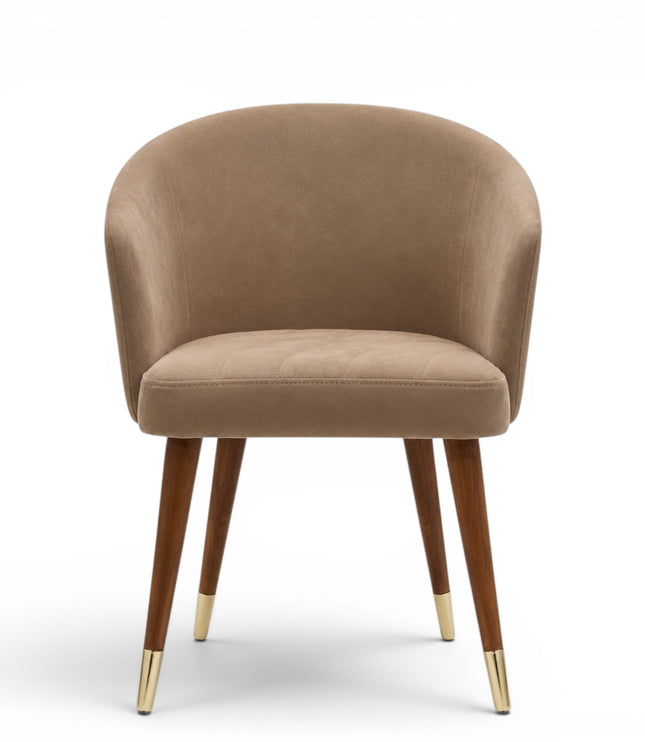 A modern dining chair with a curved, tufted backrest and seat upholstered in a warm taupe-beige fabric, supported by four tapered dark brown wooden legs with gold metal caps, shown against a plain white background.