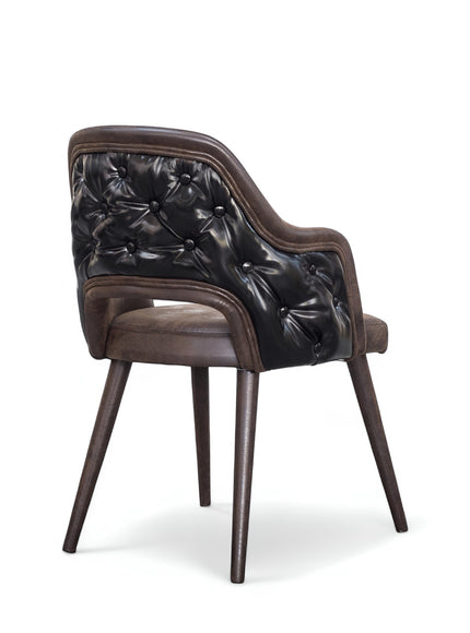 Rear angled view of the Vaya Home Alt dining chair with dark brown leather upholstery, button-tufted outer back, curved arms, and tapered wood legs.