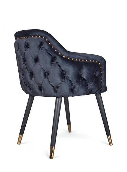 Rear angled view of the Vaya Home Lila accent chair featuring dark blue velvet upholstery, nailhead trim, tufted back detail, and black legs with gold caps.