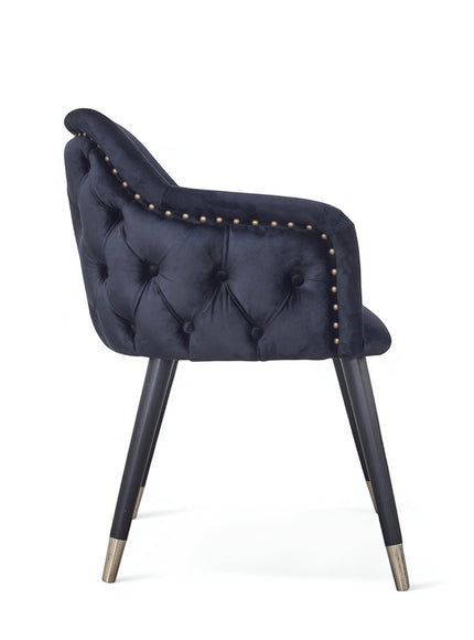 Side view of the Vaya Home Lila accent chair with dark blue velvet upholstery, button-tufted back, nailhead trim, and black legs with gold caps.