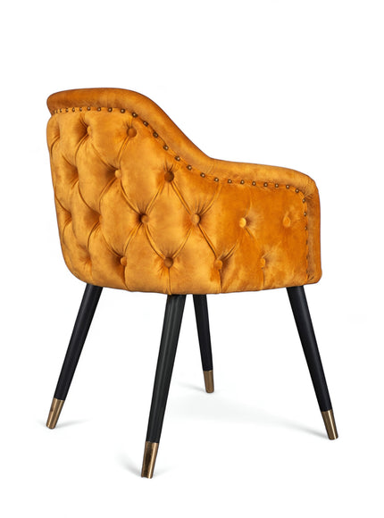 Side view of the Vaya Home Lila accent chair with orange velvet upholstery, button-tufted back, nailhead trim, and black legs with gold caps.