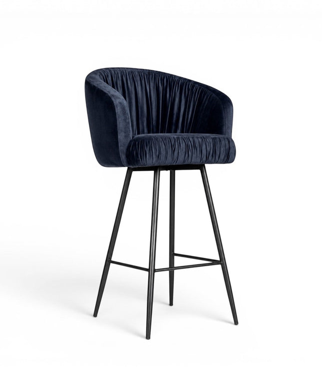 Front angled view of the Vaya Home Midnight bar stool with dark blue velvet upholstery, a channel-tufted curved back, and a black metal base.