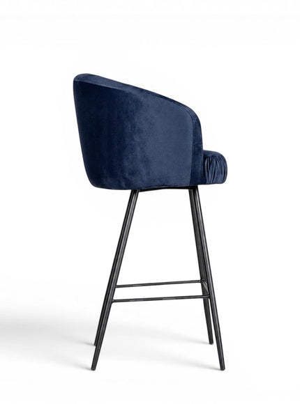 Side view of the Vaya Home Midnight bar stool featuring dark blue velvet upholstery, a curved backrest, and a black metal base.
