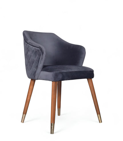 Front angled view of the Vaya Home Rioga dining chair with dark blue velvet upholstery, curved armrests, and tapered wood legs with gold caps.