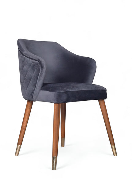 Front angled view of the Vaya Home Rioga dining chair with dark blue velvet upholstery, curved armrests, and tapered wood legs with gold caps.