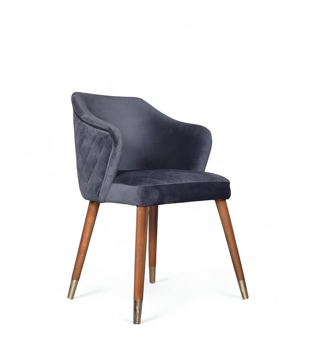 Front angled view of the Vaya Home Rioga dining chair with dark blue velvet upholstery, curved armrests, and tapered wood legs with gold caps.