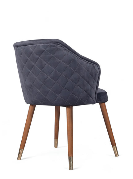 Rear angled view of the Vaya Home Rioga dining chair featuring dark blue velvet upholstery, diamond-quilted back detail, and tapered wood legs with gold caps.