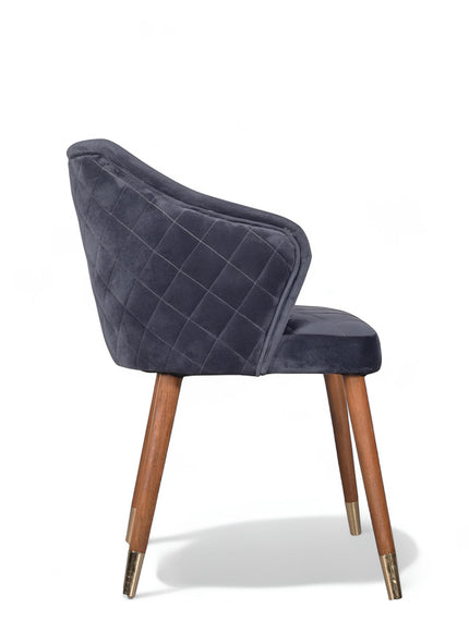 Side view of the Vaya Home Rioga dining chair with dark blue velvet upholstery, curved arms, quilted outer back, and tapered wood legs with gold caps.