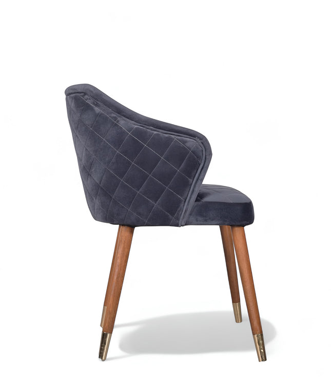 Side view of the Vaya Home Rioga dining chair with dark blue velvet upholstery, curved arms, quilted outer back, and tapered wood legs with gold caps.