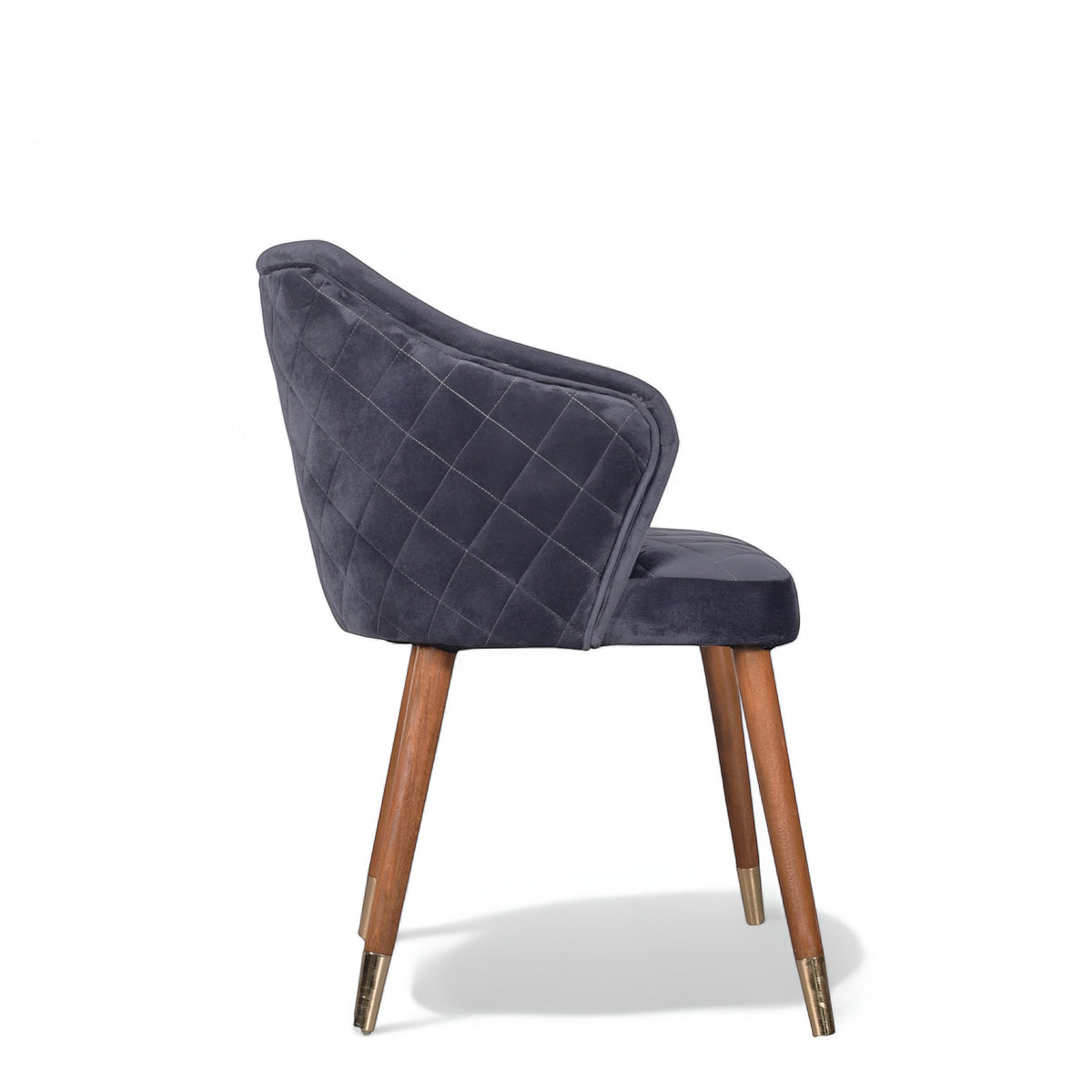 Side view of the Vaya Home Rioga dining chair with dark blue velvet upholstery, curved arms, quilted outer back, and tapered wood legs with gold caps.