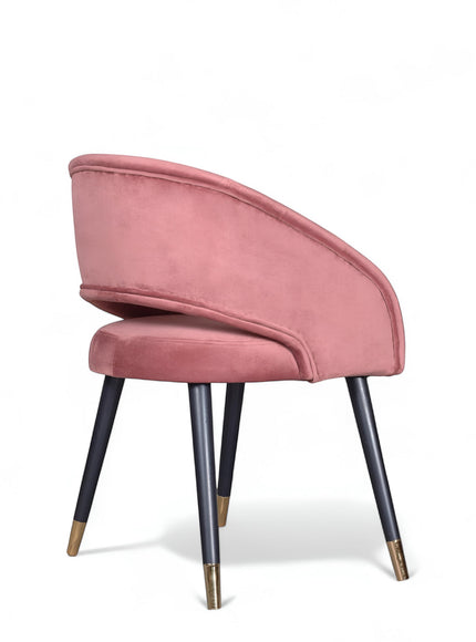 Rear angled view of the Vaya Home Rosado dining chair featuring pink velvet upholstery, a rounded back, and black tapered legs with gold caps.