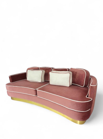 Angled view of the Vaya Home Rose curved sofa featuring pink velvet upholstery, a sculpted curved silhouette, contrast piping, and accent cushions.