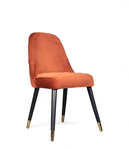 Front angled view of the Vaya Home Stretto dining chair with orange velvet upholstery, slim profile, and black tapered legs with gold caps.