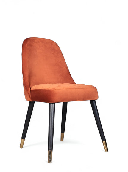 Front angled view of the Vaya Home Stretto dining chair with orange velvet upholstery, slim profile, and black tapered legs with gold caps.