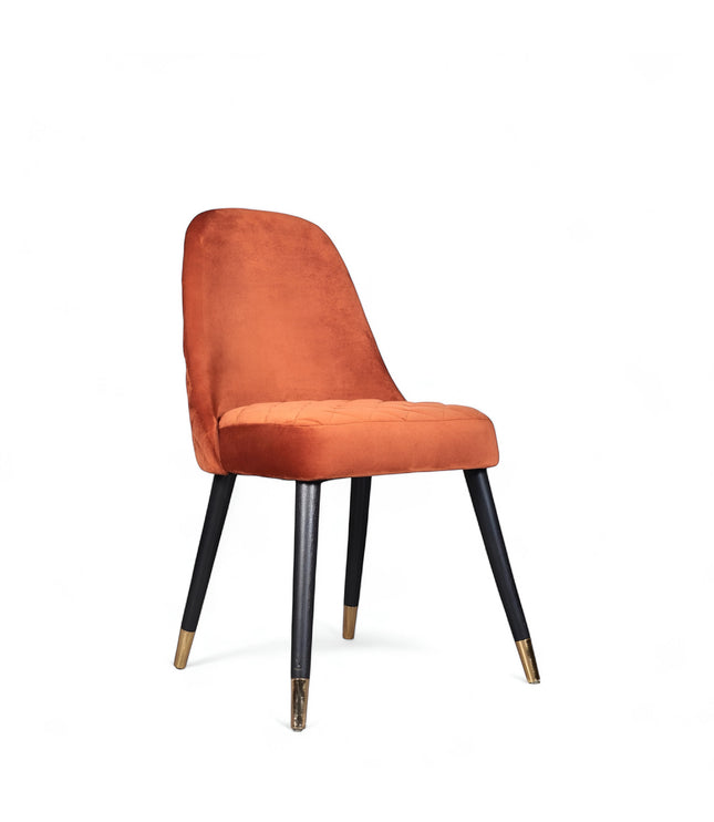Front angled view of the Vaya Home Stretto dining chair with orange velvet upholstery, slim profile, and black tapered legs with gold caps.