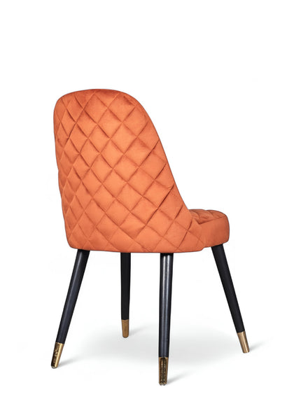 Rear angled view of the Vaya Home Stretto dining chair featuring orange velvet upholstery, diamond-quilted back detail, and black tapered legs with gold caps.