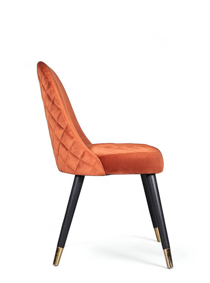 Side view of the Vaya Home Stretto dining chair with orange velvet upholstery, quilted backrest, and black tapered legs with gold caps.