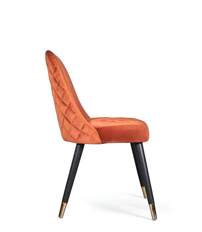 Side view of the Vaya Home Stretto dining chair with orange velvet upholstery, quilted backrest, and black tapered legs with gold caps.
