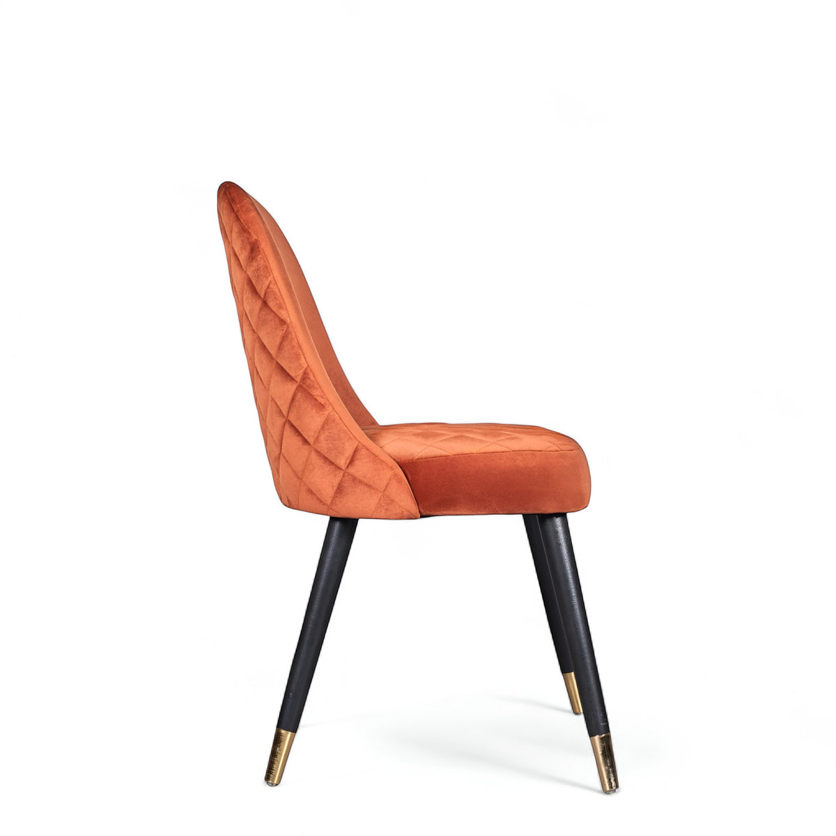 Side view of the Vaya Home Stretto dining chair with orange velvet upholstery, quilted backrest, and black tapered legs with gold caps.